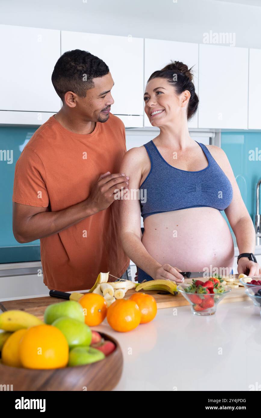 A casa, coppia multirazziale, preparando insalata di frutta sana, donna incinta e partner in cucina Foto Stock