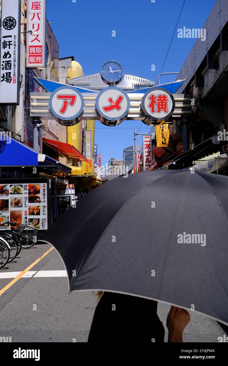 Una persona con un ombrello con il cancello d'ingresso della strada dello shopping Ameyoko sullo sfondo in una calda giornata estiva. Ueno, quartiere commerciale di Okachimachi, Taito Ward, Tokyo, Giappone Foto Stock