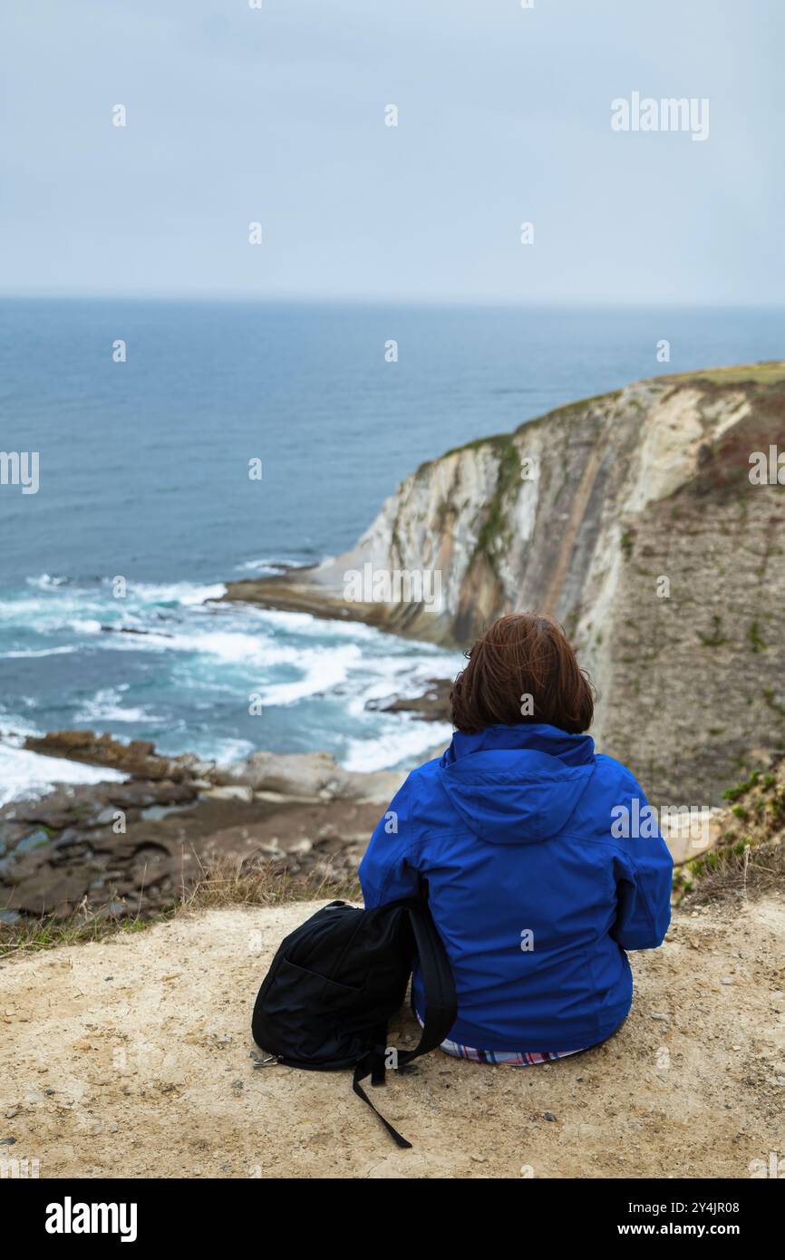 Donna caucasica in giacca blu seduta sulle scogliere che si affacciano sul mare, Paesi Baschi, Spagna. Foto Stock