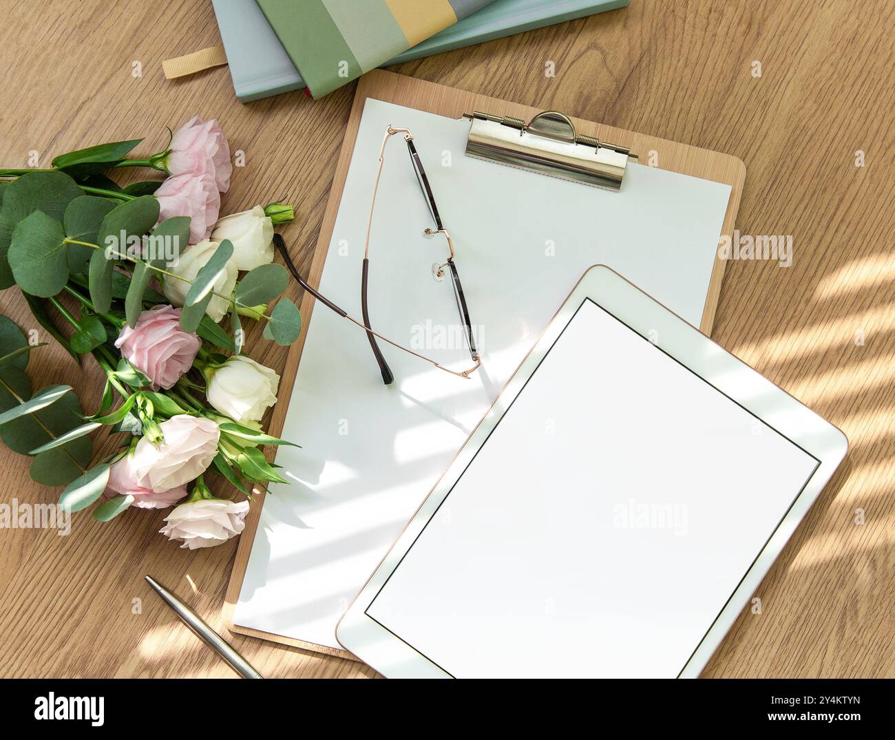 Uno spazio di lavoro luminoso è dotato di un tablet vuoto, un notebook aperto, utensili di cancelleria e fiori freschi disposti su un tavolo di legno, invitando la creatività e. Foto Stock