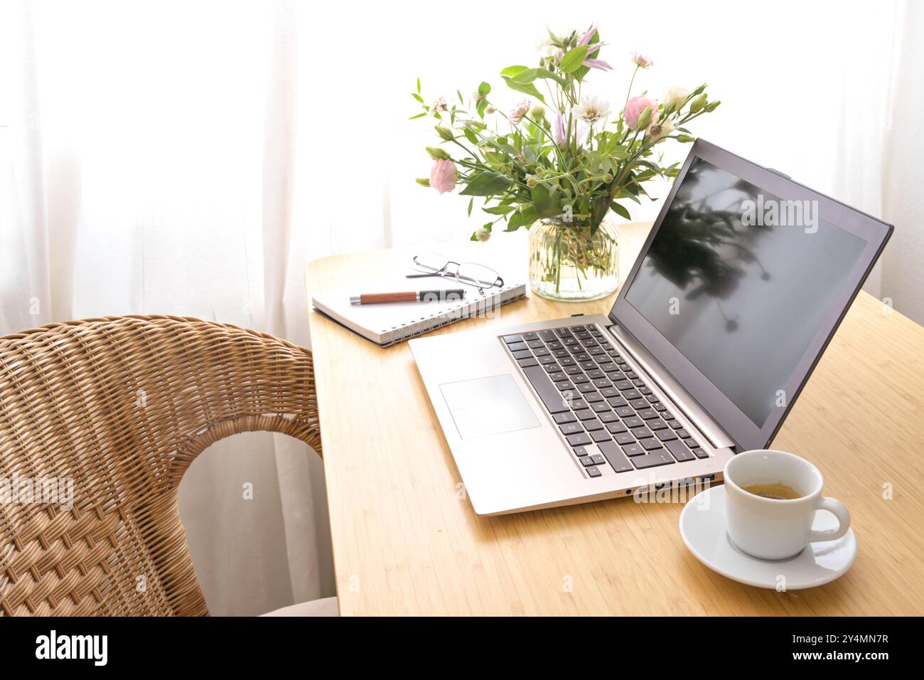 Computer portatile, caffè, bouquet di fiori e cancelleria su un tavolo di legno con poltrona in vimini, in un ufficio domestico caldo e accogliente, creativo Foto Stock