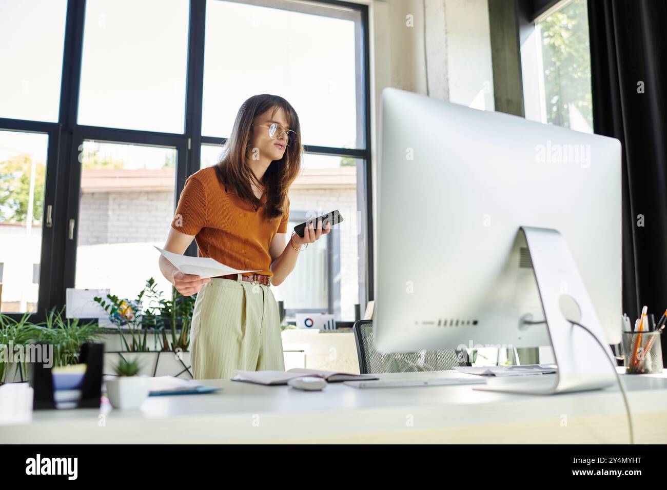 Una giovane persona non binaria bilancia i documenti e un telefono, impegnata in un lavoro d'ufficio produttivo. Foto Stock
