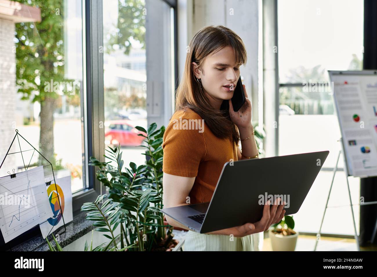 Il giovane individuo non binario al telefono, bilanciando le attività lavorative in un ufficio luminoso. Foto Stock