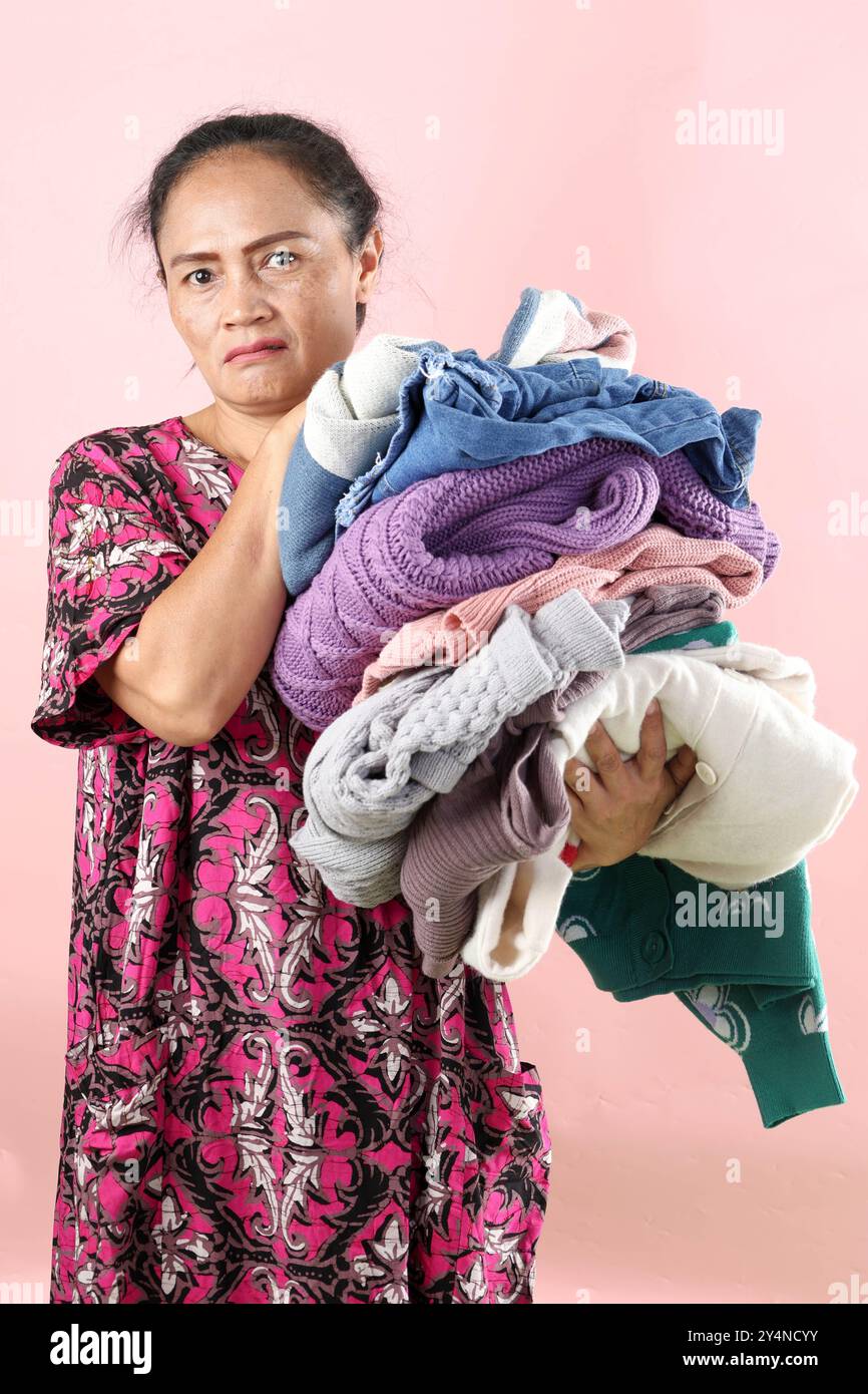 Mamma asiatica che tesa Daster che tiene vestiti ruvidi lavanderia con espressione del viso arrabbiato, su sfondo rosa Foto Stock