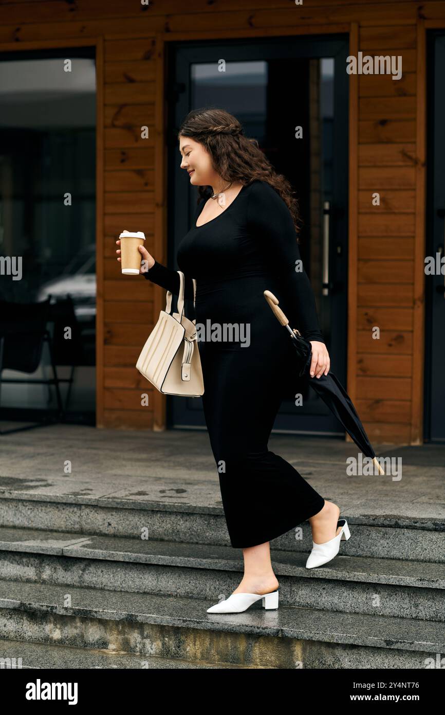 Una giovane donna passeggia all'aperto con un elegante abito nero, godendosi una giornata autunnale con una bevanda calda in mano. Foto Stock