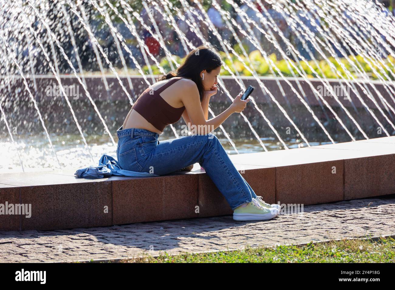 Ragazza attraente che usa lo smartphone seduto sulla strada della città sullo sfondo di una fontana Foto Stock