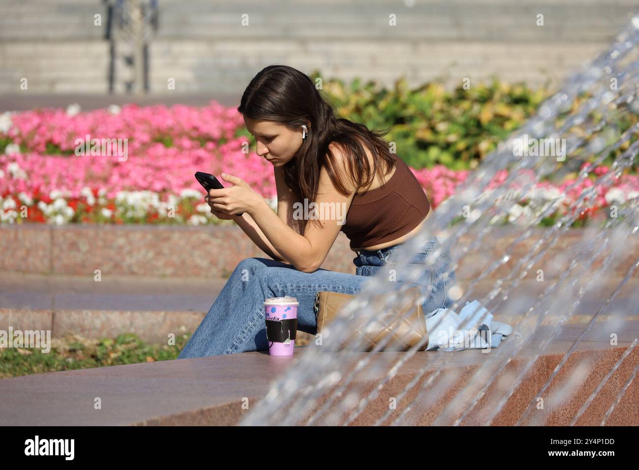 Ragazza attraente che usa lo smartphone seduto sulla strada della città sullo sfondo di una fontana Foto Stock