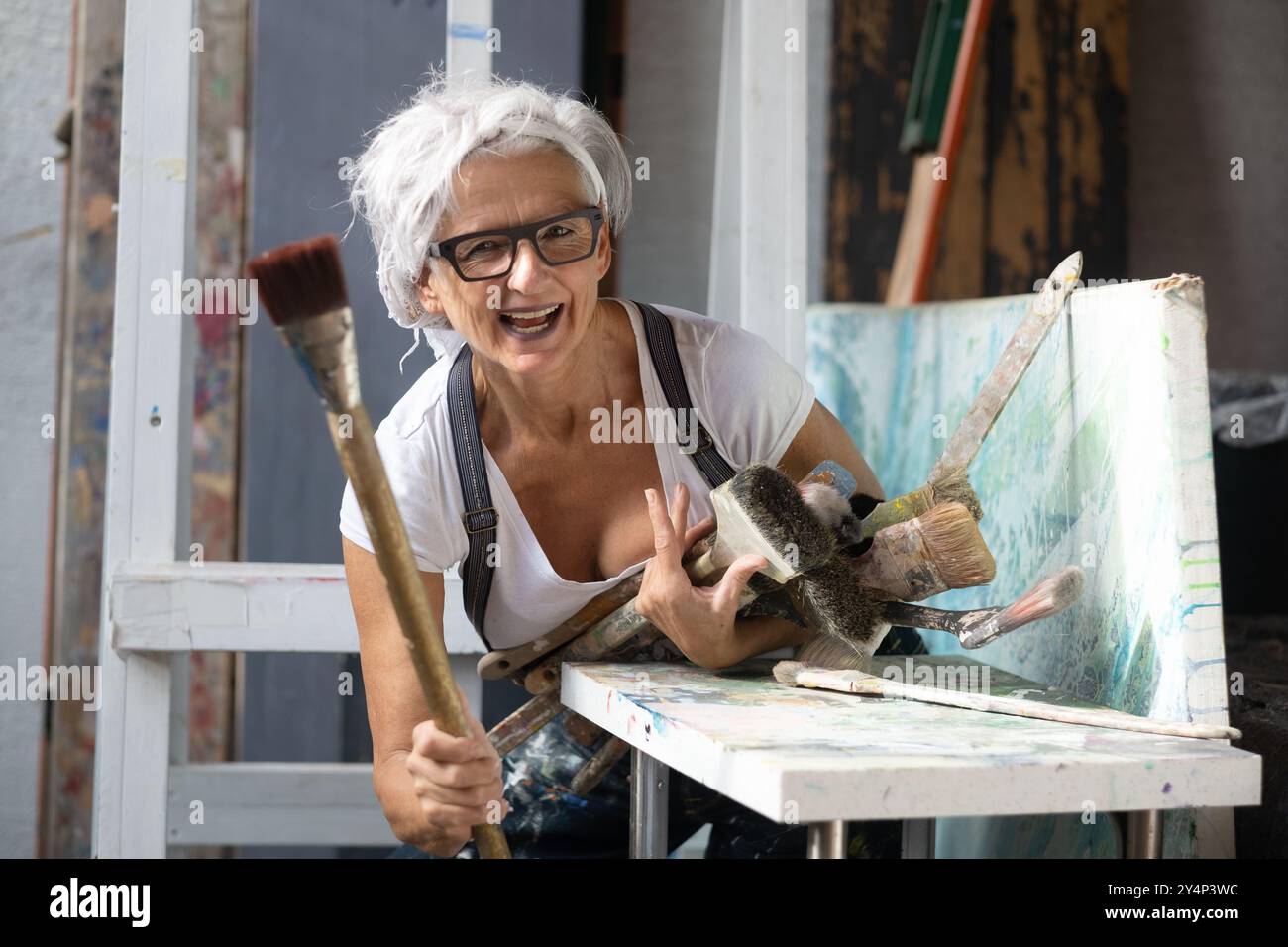 artista donna anziana che sorride energicamente nel suo studio all'aperto, tenendo diversi pennelli, circondati da tele. La sua passione per la creatività e.. Foto Stock