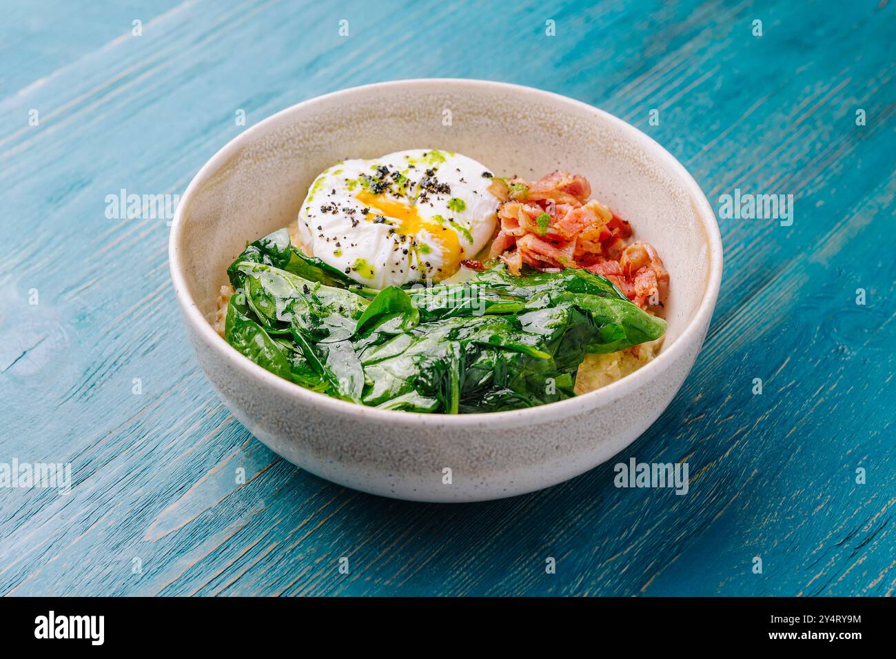 Nutriente ciotola per colazione con uovo in camicia, verdure selvagge e pancetta croccante su un rustico tavolo blu Foto Stock