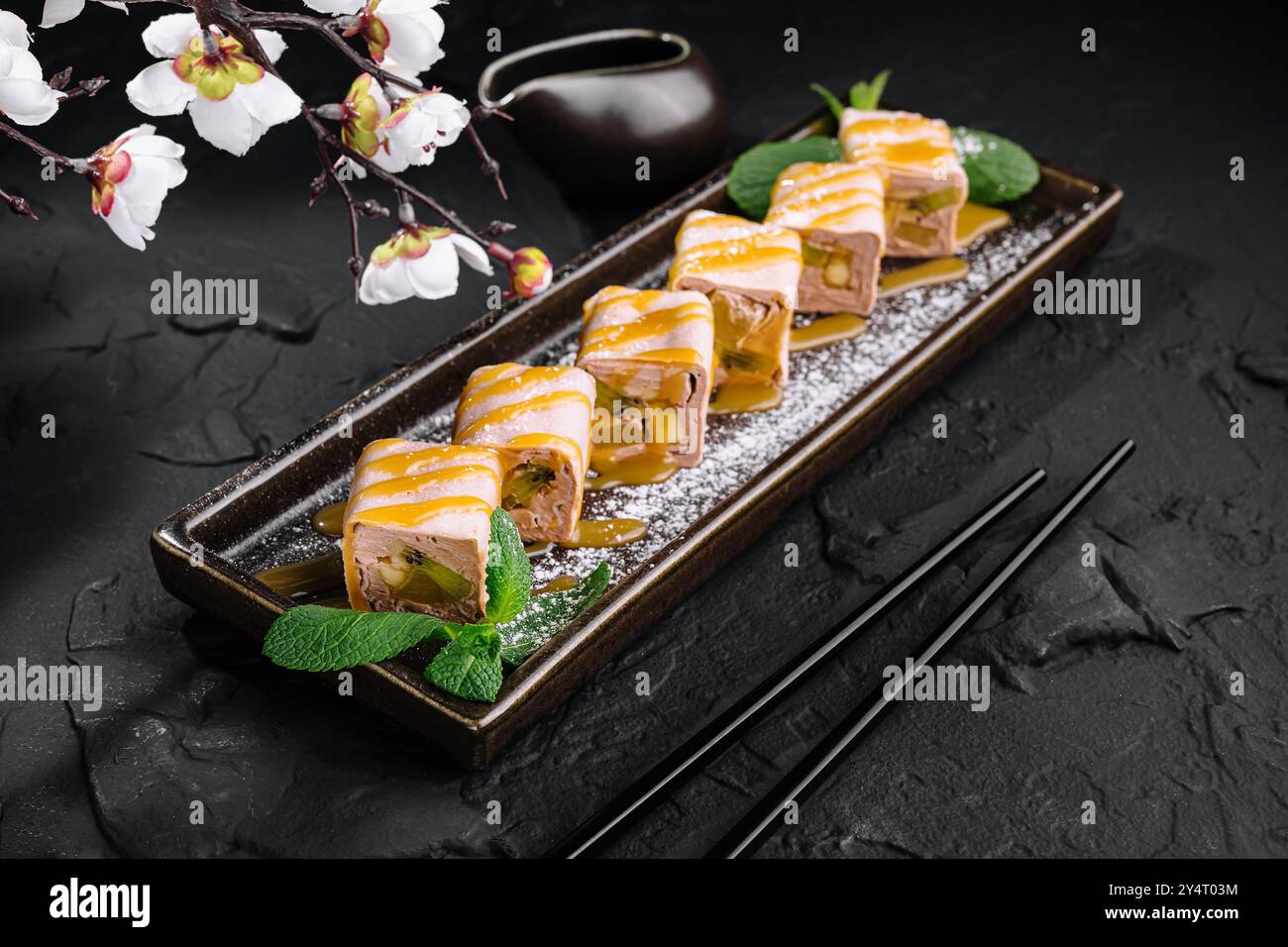 Eleganti sushi Rolls con kiwi e banana serviti su un piatto con un ramo sakura, su uno sfondo di pietra scura Foto Stock