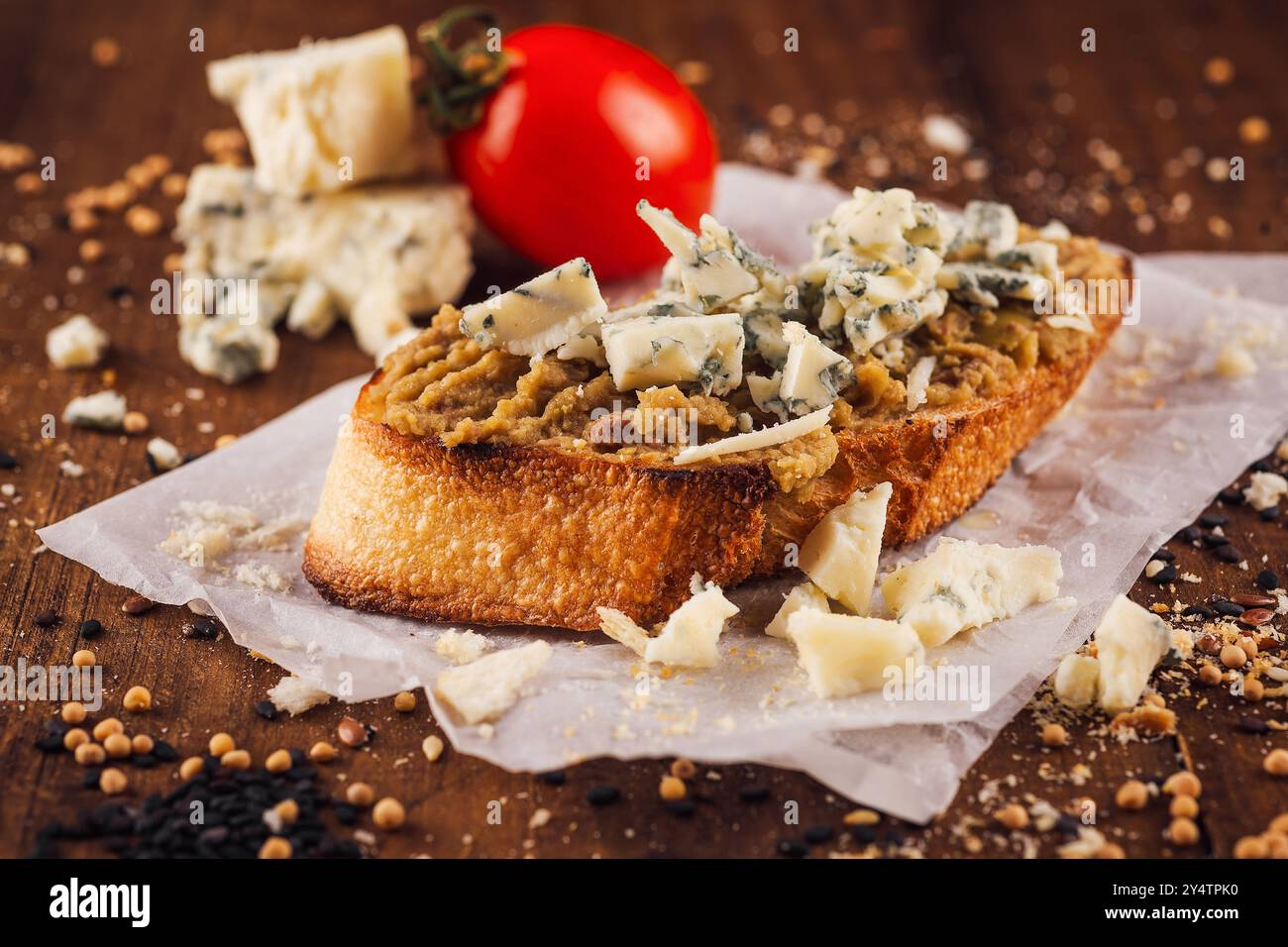 La baguette tostata adagiata su carta bianca è ricoperta di pate cremoso, cosparsa di formaggio blu e decorata con pomodoro Foto Stock