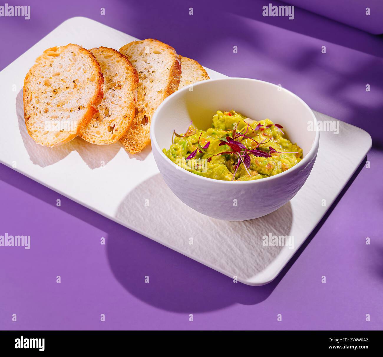 Ciotola di guacamole con fiori viola e fette di pane tostato su un piatto bianco su sfondo viola Foto Stock