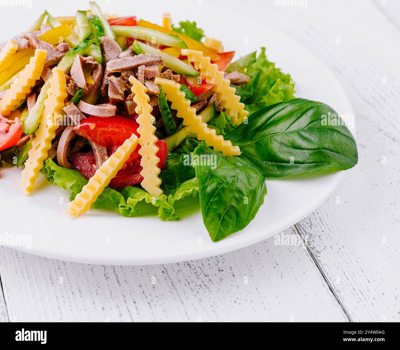 Deliziosa insalata con carne, pomodoro, cetrioli, patatine al formaggio e basilico su piatto bianco su sfondo bianco in legno Foto Stock