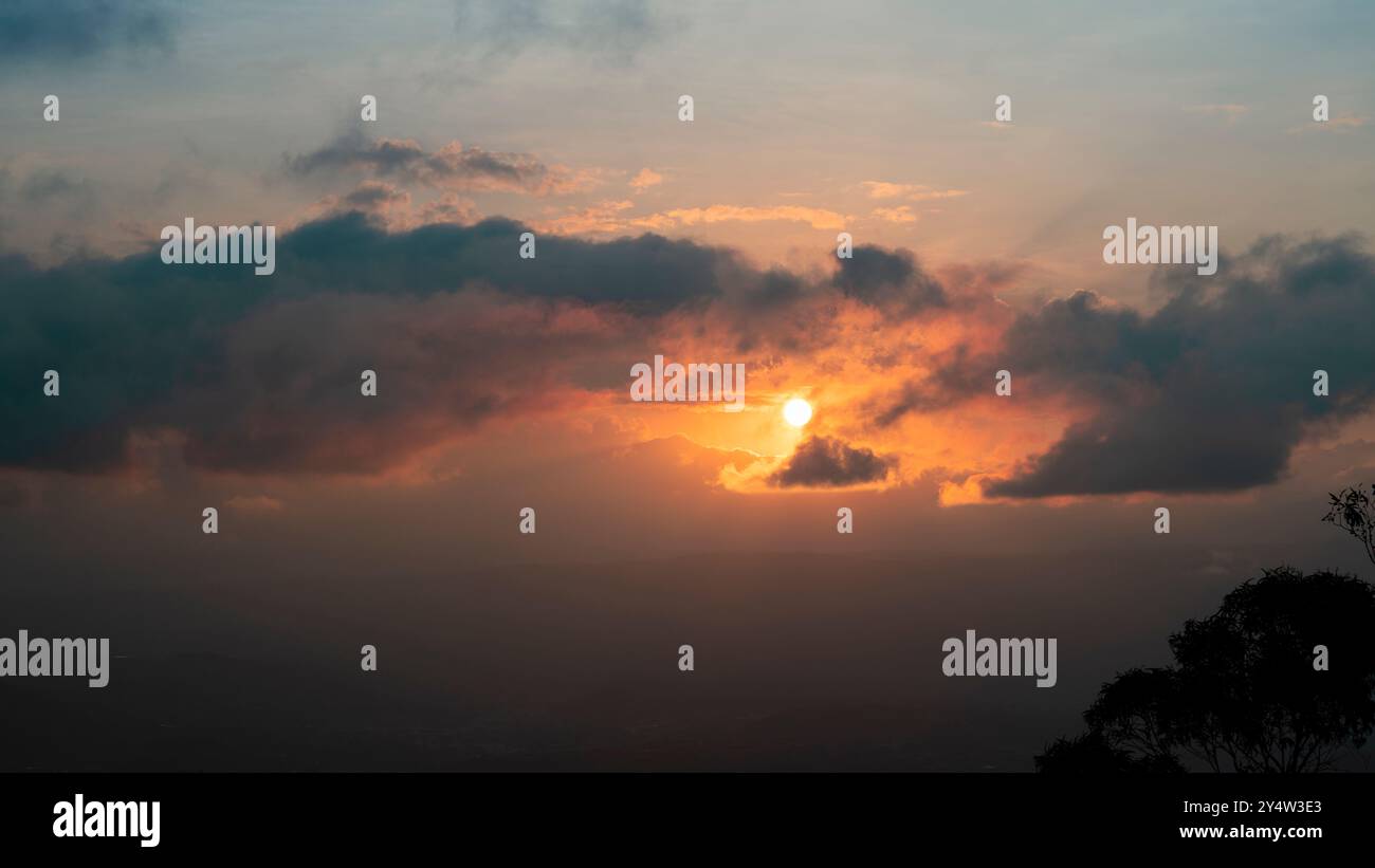 Vista panoramica del tramonto con il sole che tramonta tra le nuvole con sagome nere di alberi Foto Stock
