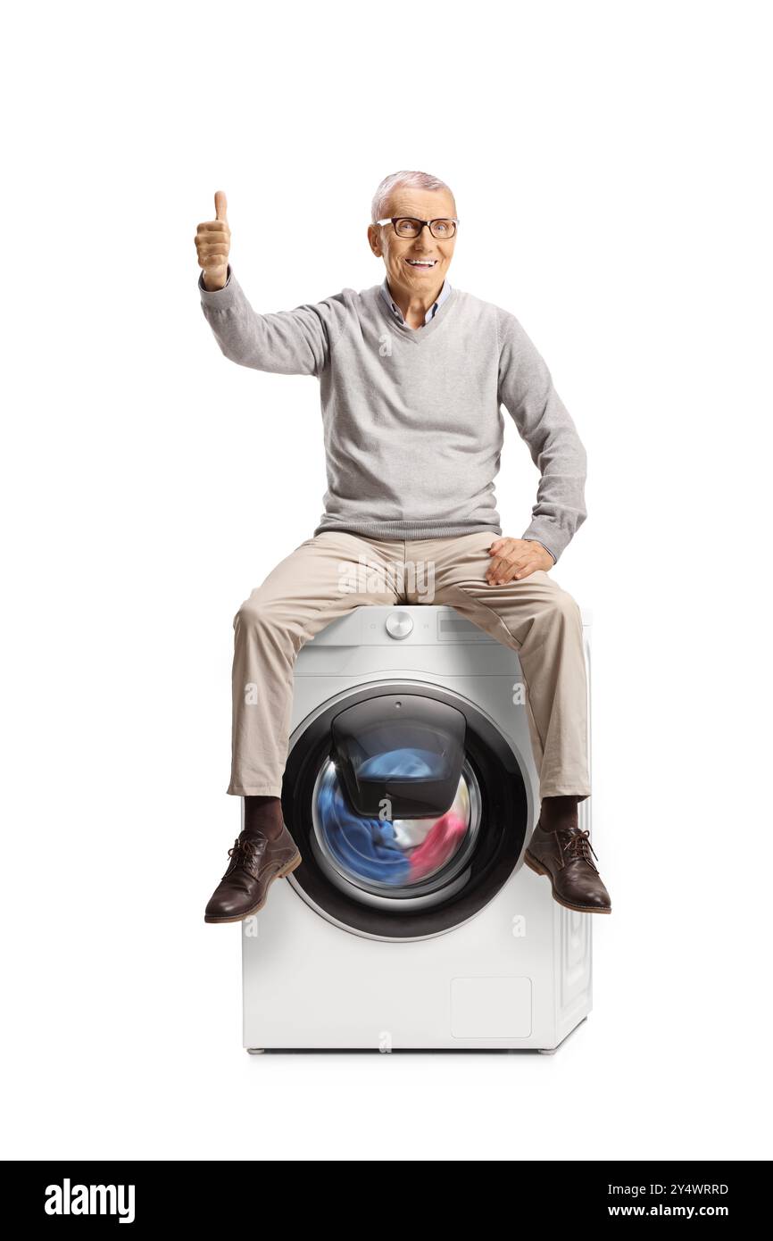 Uomo anziano felice seduto sopra una lavatrice e con gesti su sfondo bianco Foto Stock