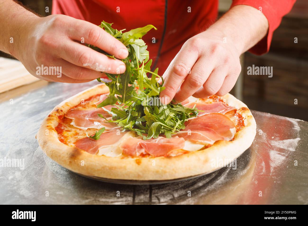 Uno chef aggiunge rucola fresca ad una pizza con prosciutto su una superficie infarinata. Foto Stock