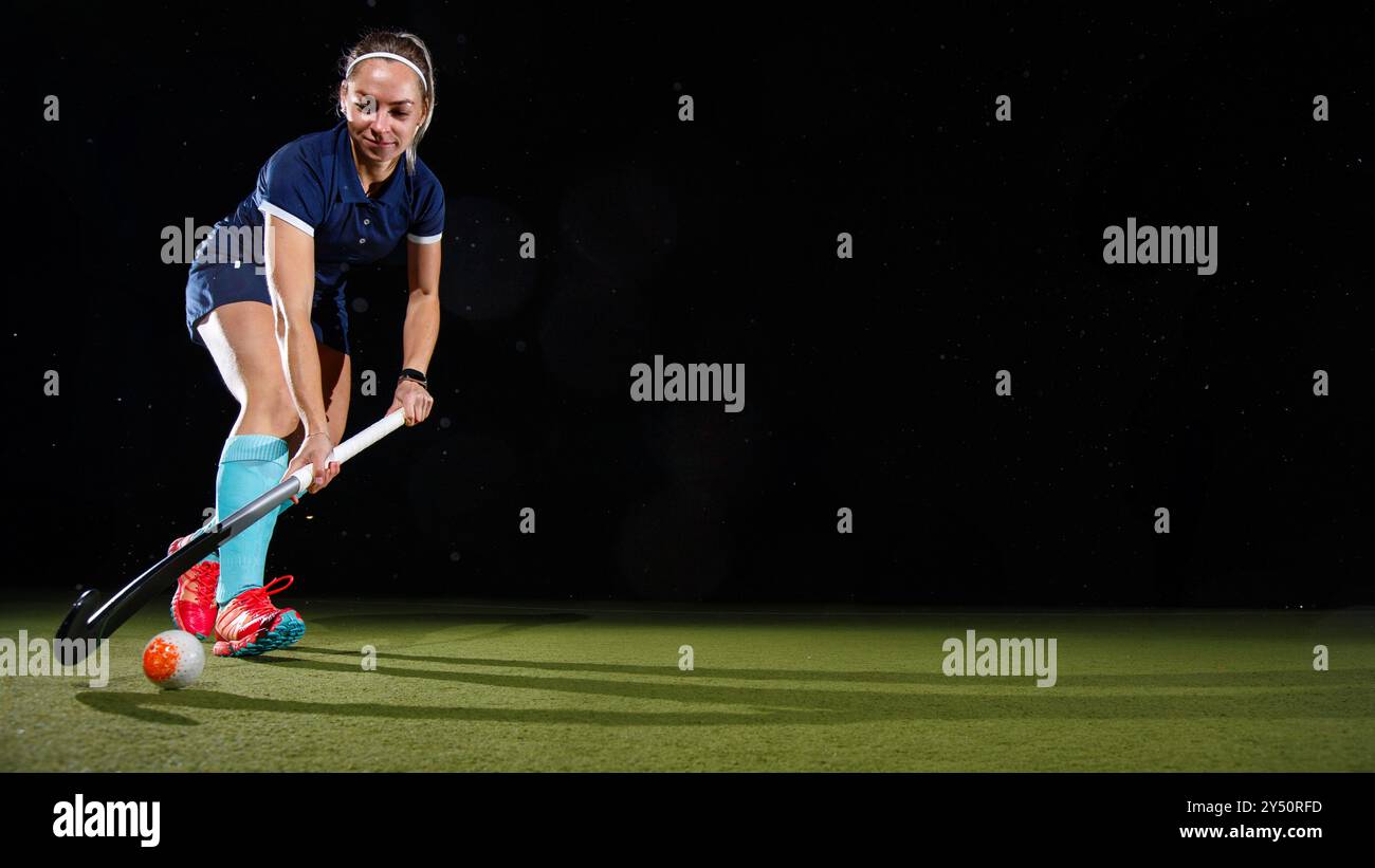 Giovane donna determinata che guida sapientemente la palla con il bastone da hockey su prato concentrato sul suo gioco. Isolato su sfondo nero Foto Stock