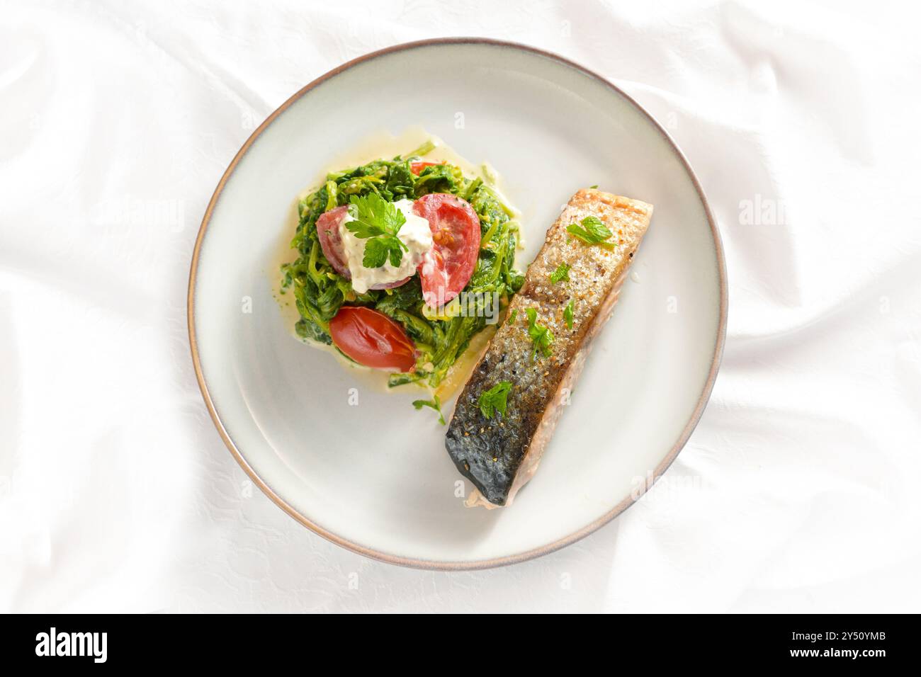 Salmone arrostito con pelle, verdure di spinaci e pomodori con crema e guarnigione di prezzemolo, sana cena a base di pesce su una tovaglia bianca, angolo alto V Foto Stock