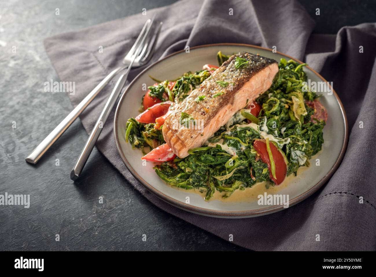 Filetto di salmone arrostito con una pelle croccante su crema di spinaci verdure con cipolle e pomodori, piatto di pesce sano con posate su un tovagliolo grigio scuro Foto Stock