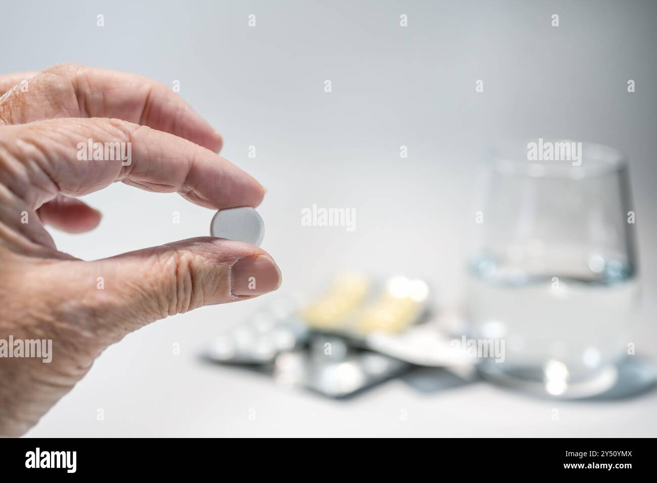 Mano anziana che tiene una pillola davanti a pacchetti di farmaci sfocati e un bicchiere d'acqua, medicina e assistenza sanitaria concetto, sfondo grigio chiaro, copia Foto Stock
