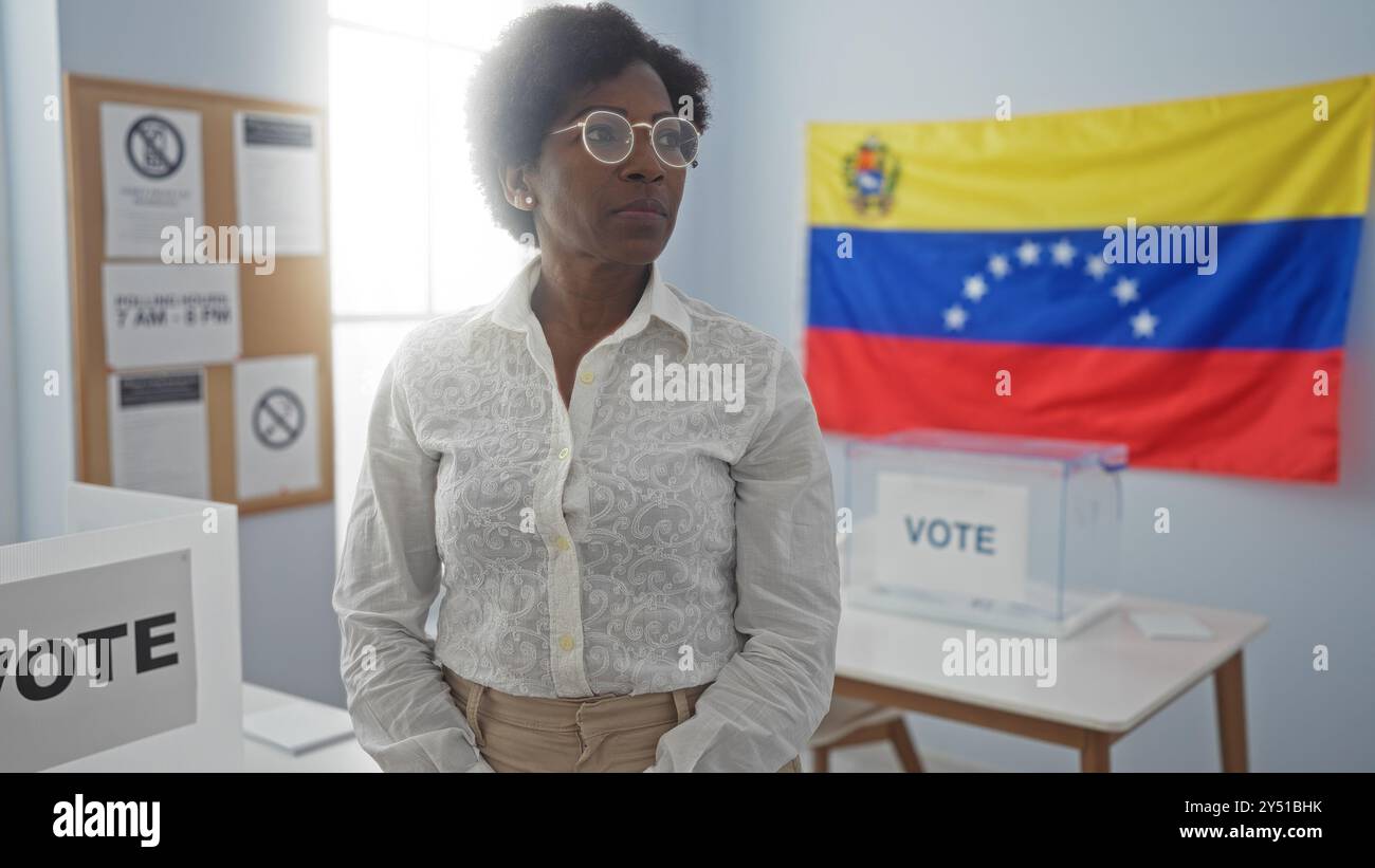 La donna afroamericana con i capelli ricci si trova in una sala del college elettorale al coperto di fronte a una bandiera venezuelana, evidenziando un processo di voto. Foto Stock