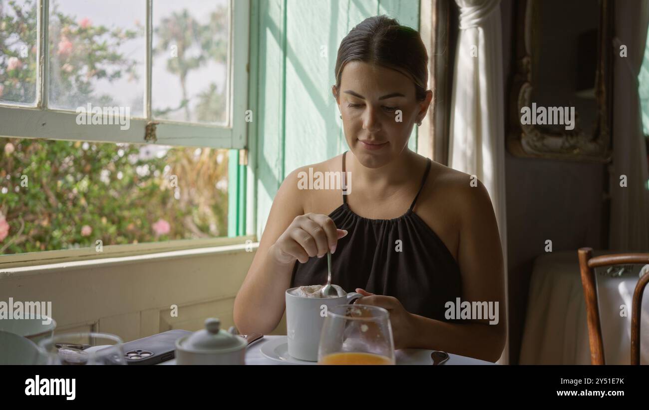 Donna che si gusta un caffè all'interno al tavolo del ristorante durante il giorno, la luce del sole scorre attraverso le finestre, circondata dal verde Foto Stock