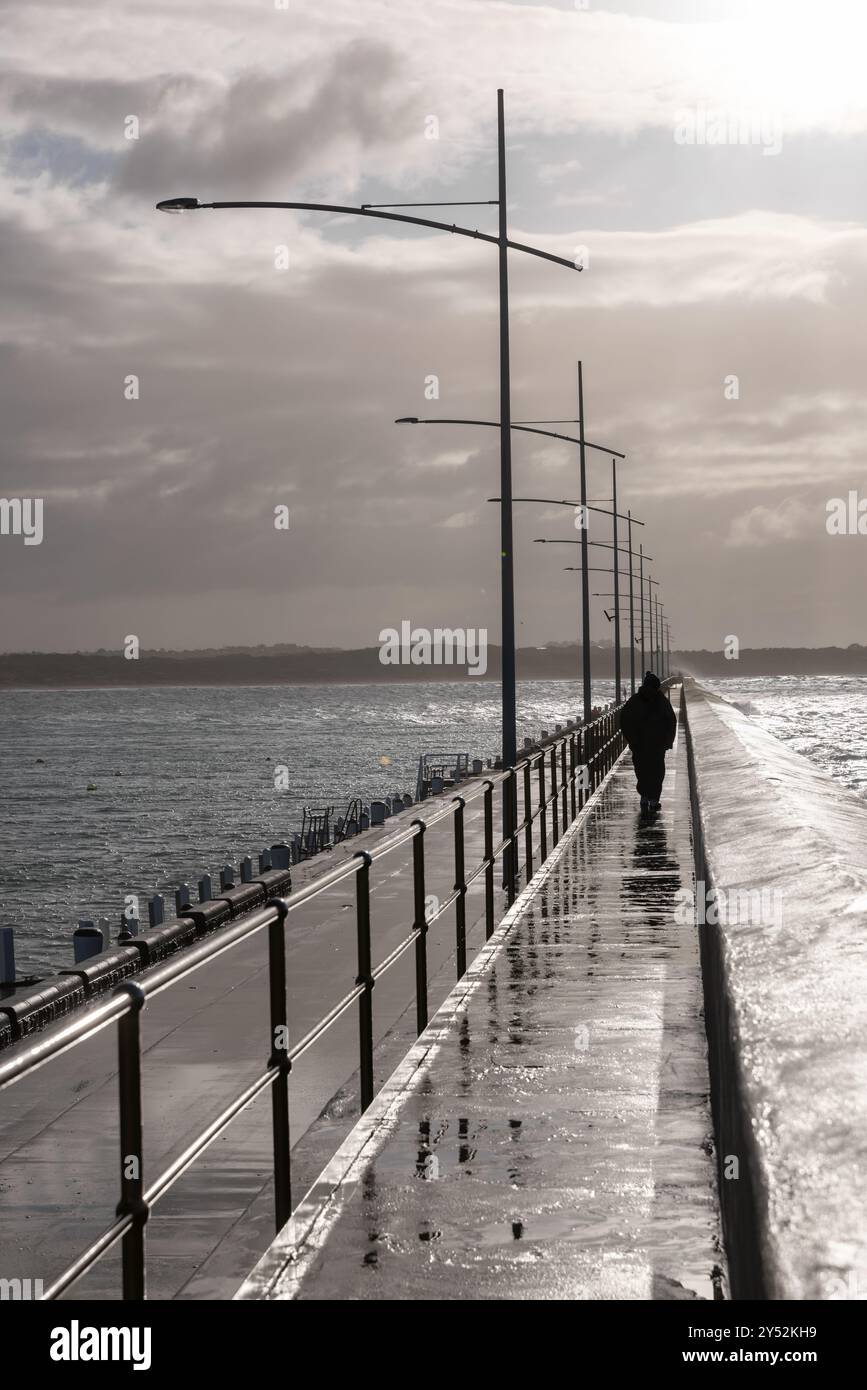 Uomo solitario che cammina su un molo bagnato dopo la pioggia, con cieli nuvolosi Foto Stock