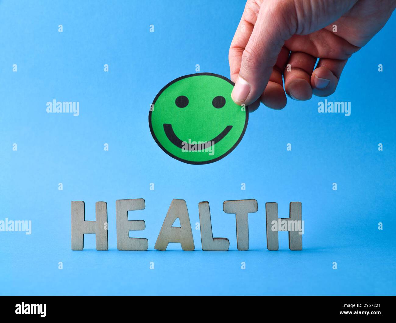 Sfondo sanitario generico con lettere ritagliate in legno e mano che tiene verde emoticon sorridente isolato su sfondo blu. Vista frontale Foto Stock