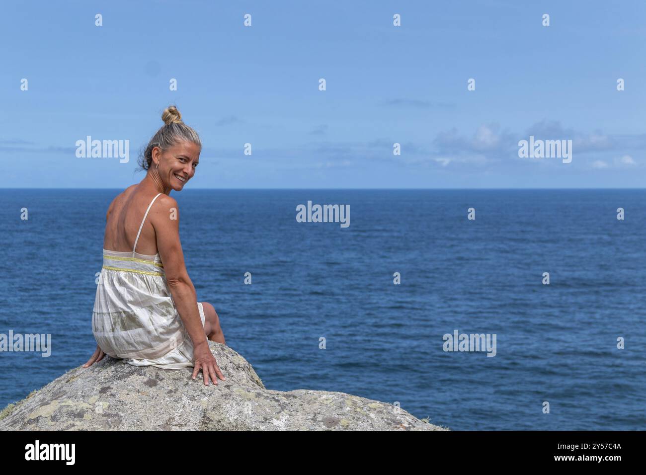 Donna bionda con i capelli in alto e indossa un abito bianco su una scogliera vicino al mare, facendo gesti con le mani. Copia spazio Foto Stock