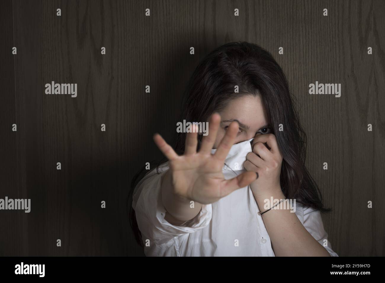 Giovane donna che nasconde il suo volto con la camicia mentre allunga la mano verso la telecamera per non essere vista, su uno sfondo di legno, nell'ombra Foto Stock