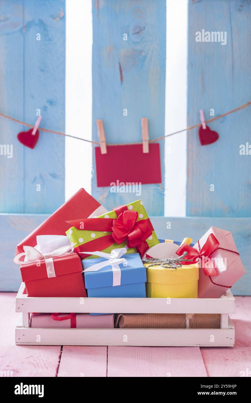 Scatole regalo colorate in una cassa di legno bianca su un tavolo rosa con una nota di carta rossa e cuori legati a una corda, su una recinzione blu Foto Stock