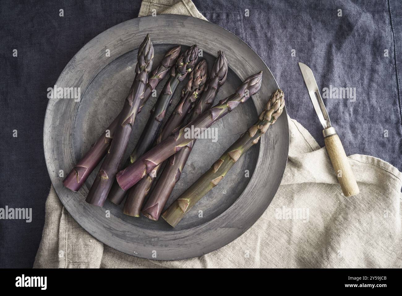 Asparagi viola freschi su un vecchio piatto di peltro con un vecchio coltello da cucina su lino scuro Foto Stock