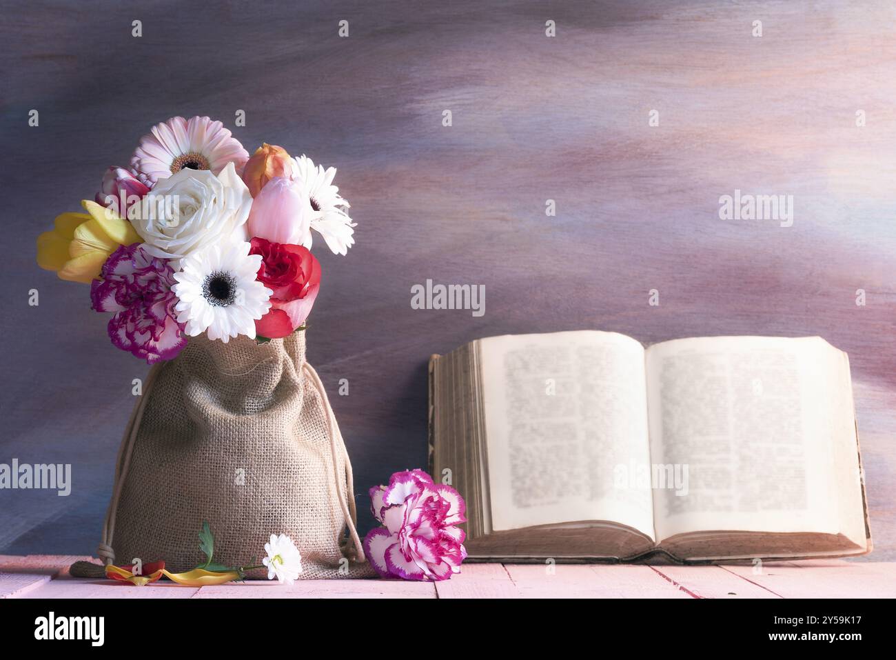 Splendido bouquet di rose, gerbera, tulipani e garofani su un tavolo rosa e un antico libro aperto appoggiato su uno sfondo di legno viola Foto Stock