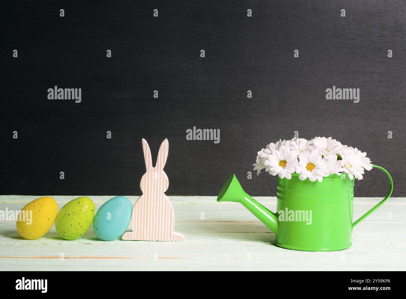 Uova dipinte di Pasqua appoggiate su un coniglietto di legno e una lattina da irrigazione verde piena di margherite bianche, su un tavolo, con una parete nera, in una giornata di sole Foto Stock