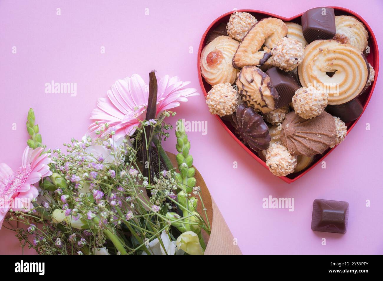 Regalare un'immagine a tema con una scatola a forma di cuore rossa piena di deliziosi dolci e un bouquet di fiori su sfondo rosa. Un'idea di biglietto d'auguri Foto Stock