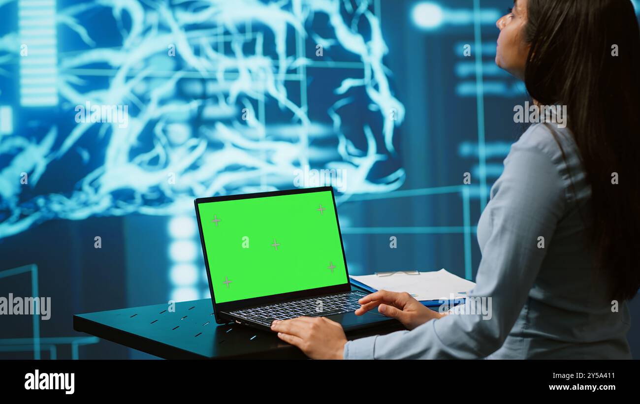 Dipendente all'avvio che guarda i grafici, utilizza il software di intelligenza artificiale su un notebook con schermo verde per prendere decisioni basate sui dati. Donna indiana che sviluppa soluzioni innovative di intelligenza artificiale per l'azienda, telecamera A Foto Stock