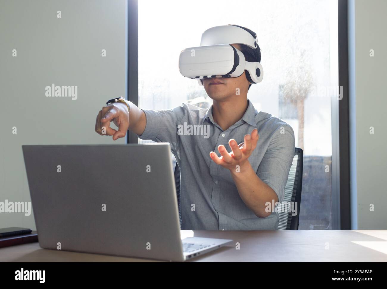 In ufficio, utilizzando il visore VR, uomo asiatico che interagisce con l'ambiente virtuale mentre lavora su un laptop Foto Stock