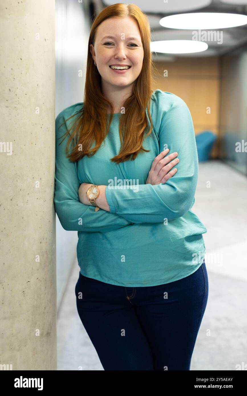 Donna sorridente in un abbigliamento casual in piedi con le braccia incrociate in ufficio Foto Stock