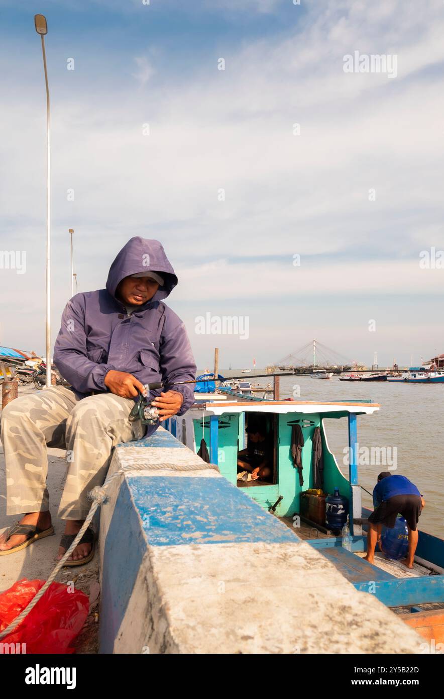Balikpapan, Borneo Orientale, Indonesia - 19 settembre. Villaggio dei pescatori di Manggar. Un uomo con una giacca blu siede su una sporgenza accanto a una barca Foto Stock