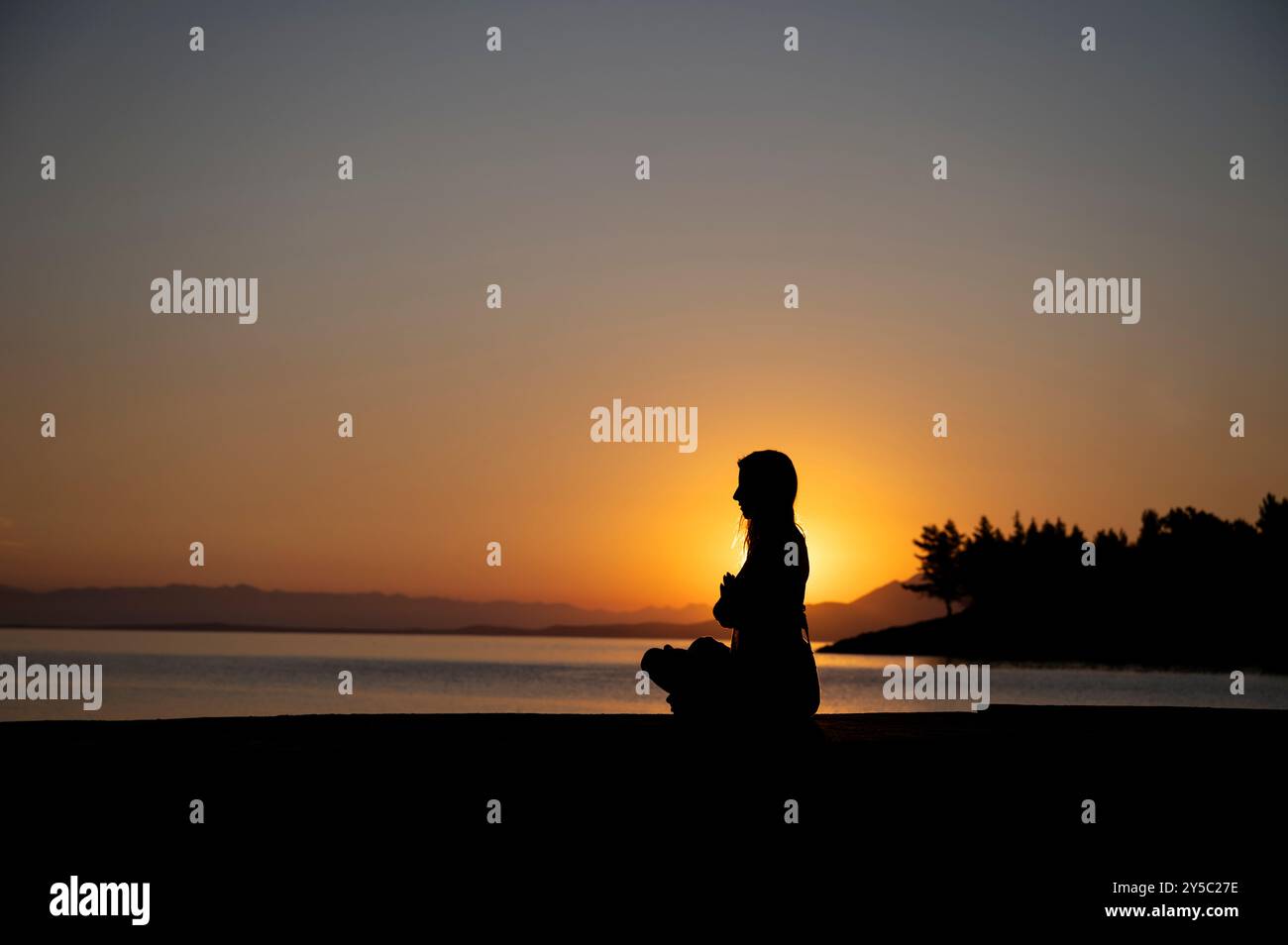 Vista del profilo di una giovane donna seduta in posizione di loto con le palme unite davanti al petto che meditava o pregava. Seduti in riva al mare al Sunri Foto Stock