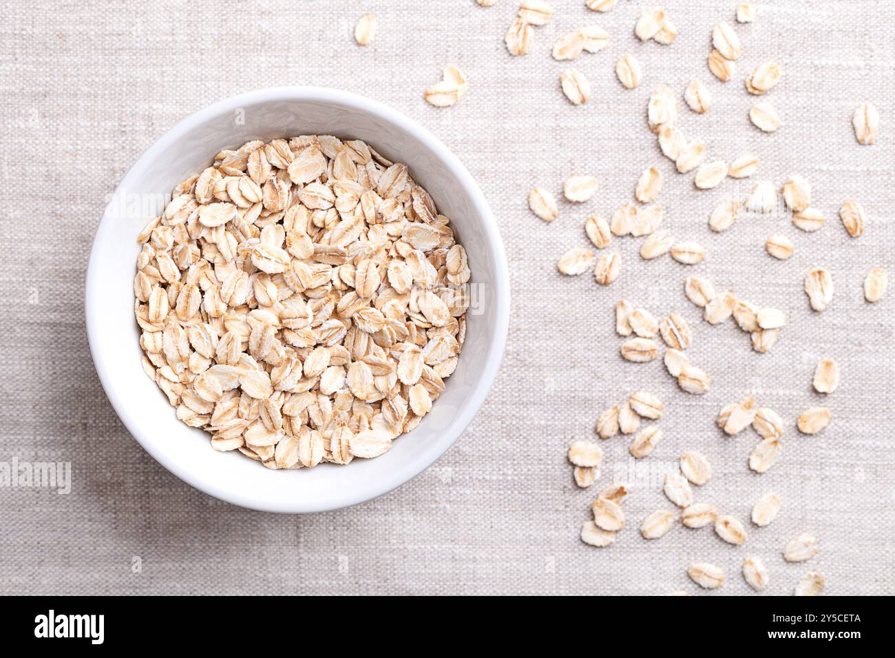 Fiocchi d'avena arrotolati, fiocchi d'avena arrotolati in una ciotola bianca su lino. Le farine d'avena cotte e cotte a vapore sono state arrotolate in fiocchi e leggermente tostate. Cibo integrale. Foto Stock