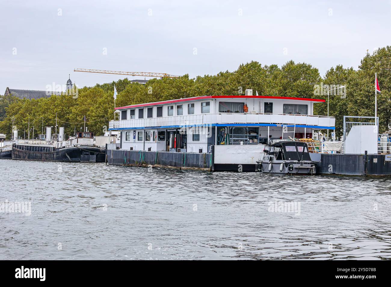 Gruppo di barche ormeggiate al molo di argine lungo il fiume Mosa, verde fogliame di alberi sullo sfondo contro il cielo grigio, acqua calma, nuvolosa giornata a Maastrich Foto Stock