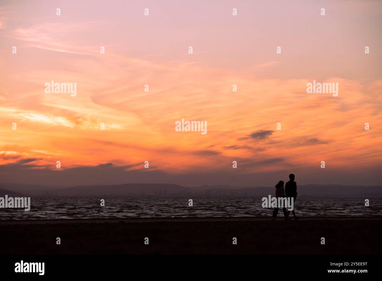 Una coppia si staglia contro il sole mentre tramonta sul Galles, come si vede da Weston-super-Mare nel Somerset. Foto Stock