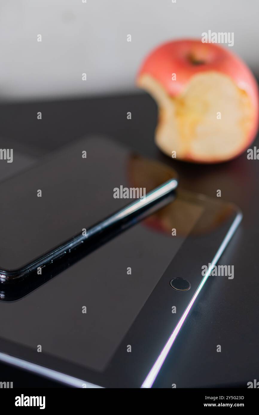 Primo piano di uno smartphone e di un tablet appoggiati su una superficie nera, con una mela rossa parzialmente mangiata sullo sfondo. I dispositivi sono dotati di schermo lucido Foto Stock