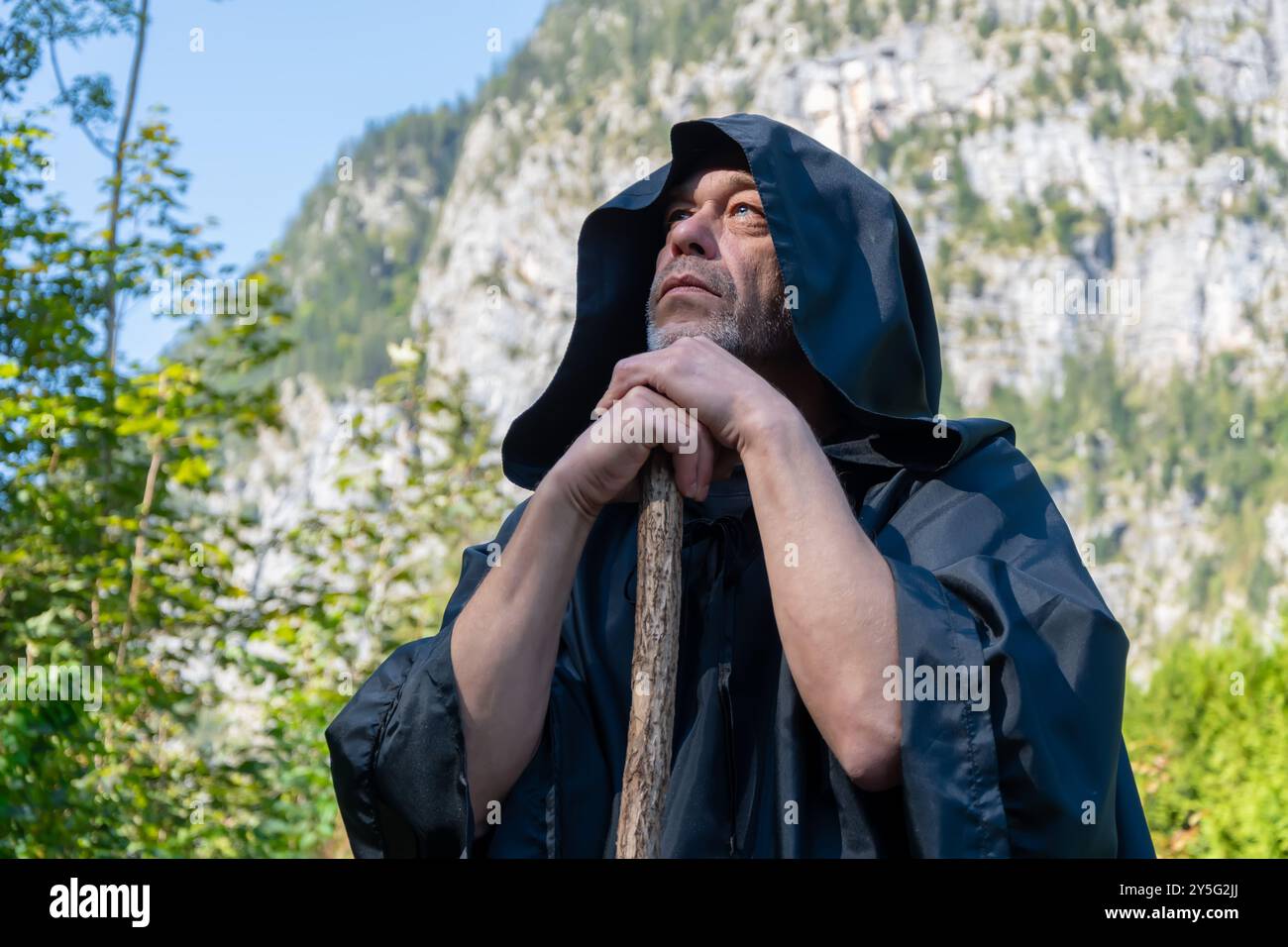 Un monaco in un mantello scuro si erge con un personale che guarda pensiamente nel cielo sullo sfondo di montagne torreggianti in una giornata di sole circondati dalla natura Foto Stock