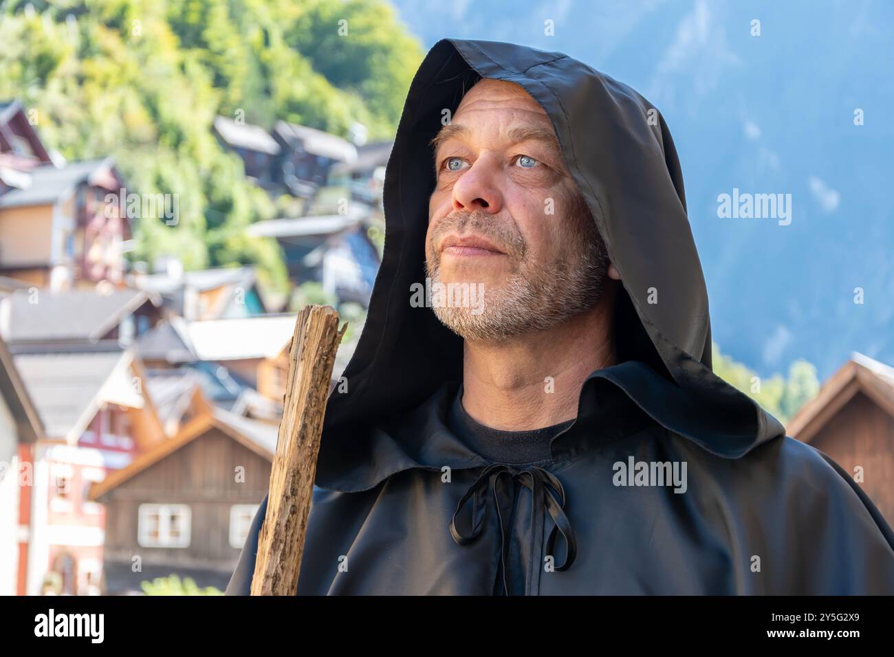 Un vecchio monaco barbuto regge un bastone di legno, affacciato sulle montagne e sul villaggio Foto Stock