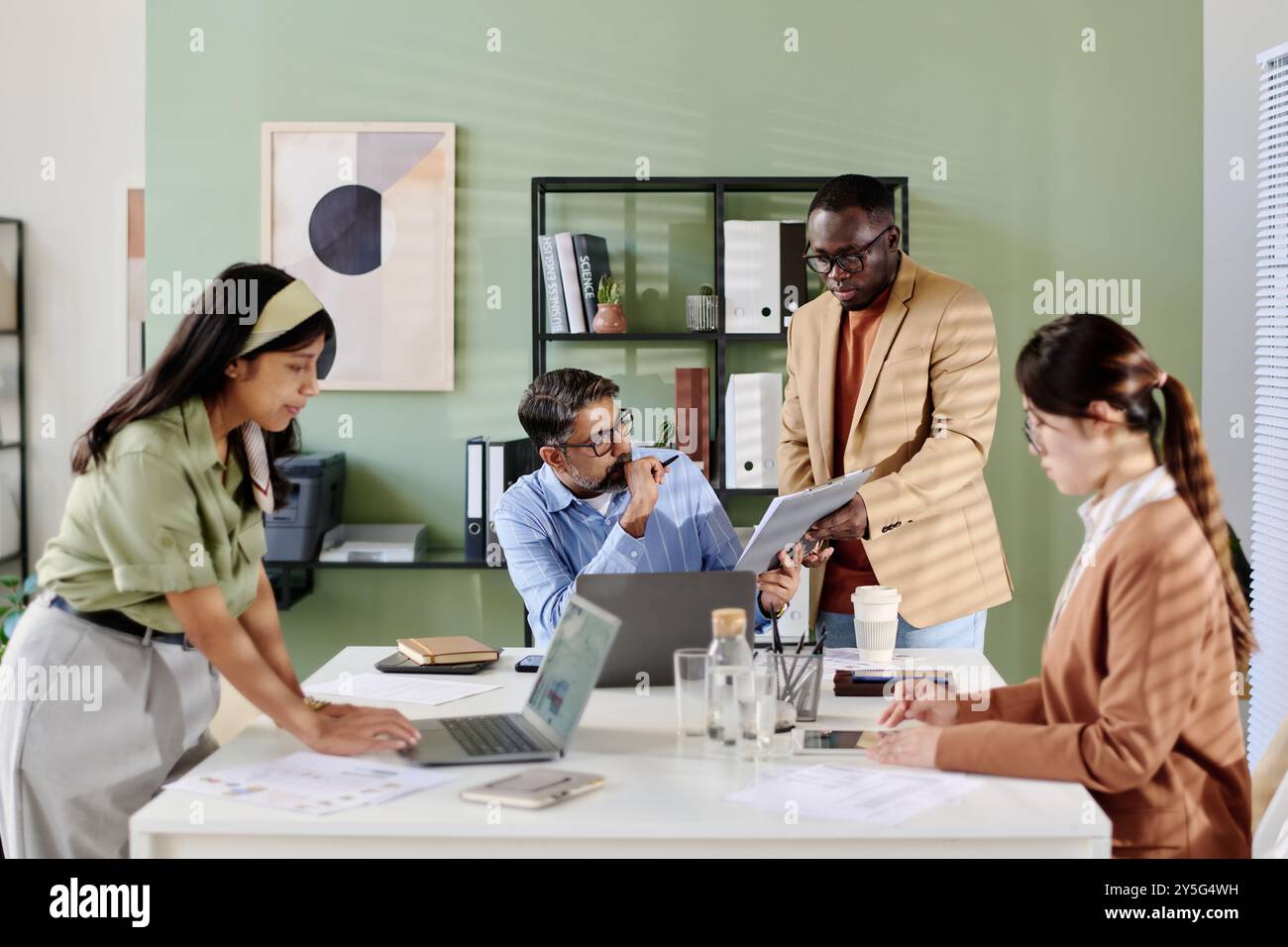 Team dell'ufficio che discute della strategia aziendale con particolare attenzione ai tablet detenuti dal membro del team in piedi. I documenti e i notebook sono sul tavolo mentre diversi team sono impegnati in discussioni Foto Stock