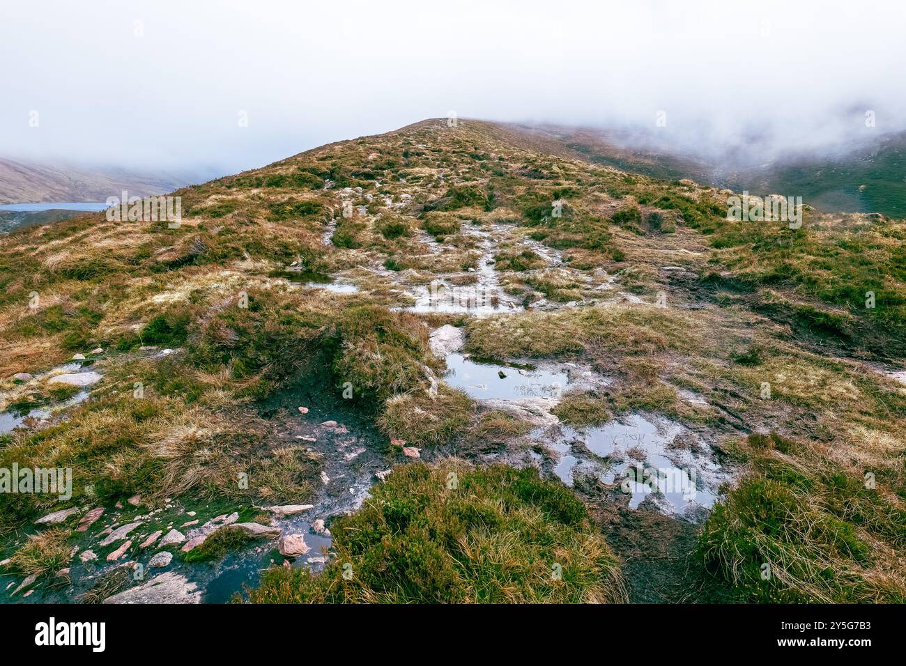 Una collina erbosa con un piccolo lago in lontananza, parzialmente oscurato da basse nuvole. Il primo piano è un sentiero fangoso con pozzanghere d'acqua. Foto Stock