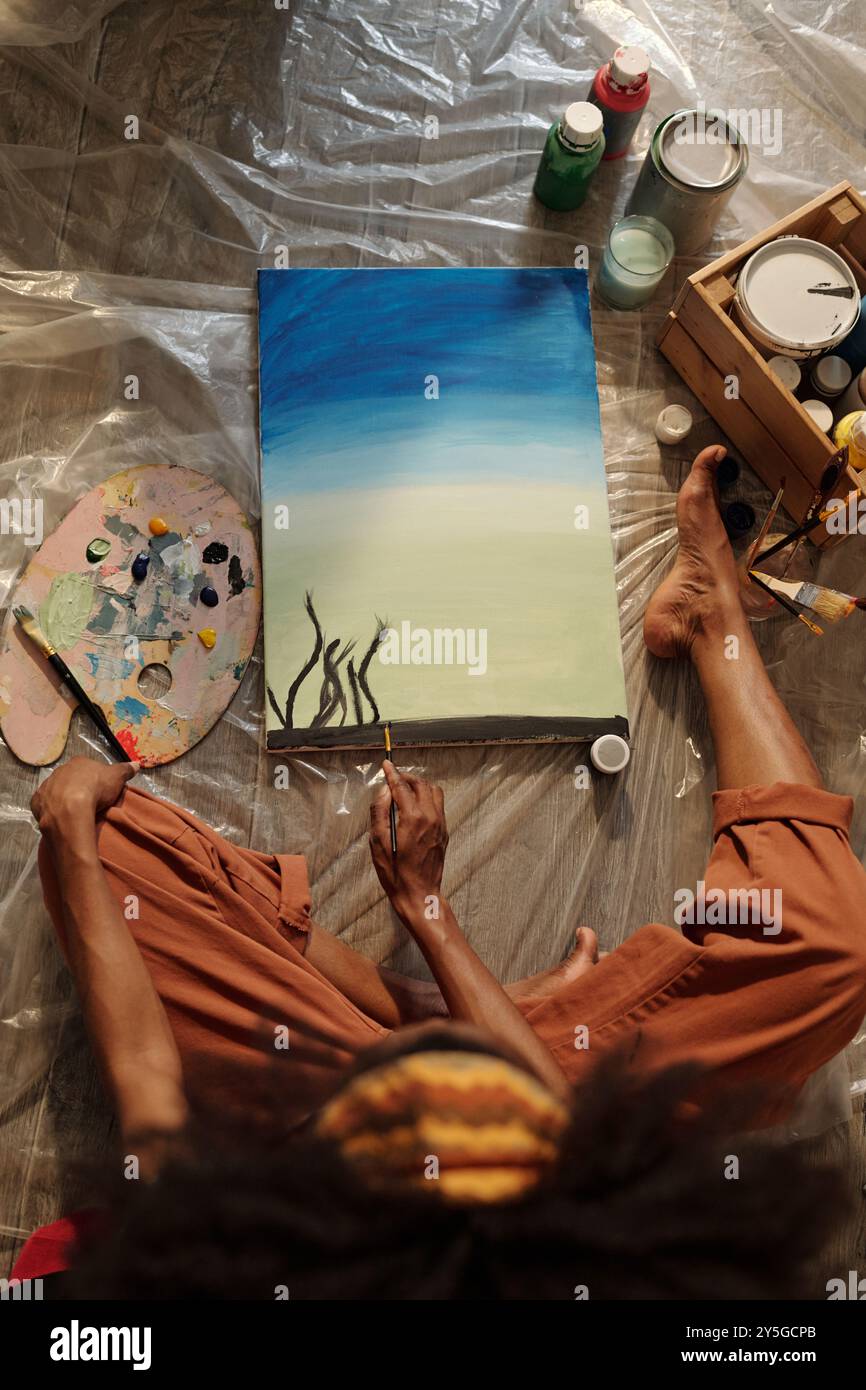 Artista seduto sul pavimento a dipingere paesaggi astratti con colori vivaci e pennelli, circondato da strumenti e materiali sparsi su lastre di plastica Foto Stock