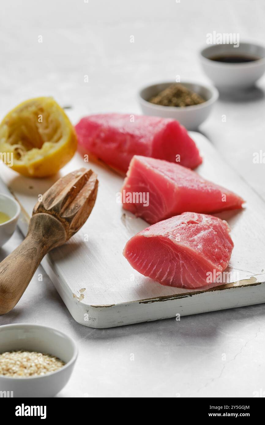 Tre vivaci bistecche di tonno riposano su un tagliere vicino alle metà del limone e alle piccole ciotole ripiene di condimento. L'allestimento suggerisce un piatto saporito nel Foto Stock
